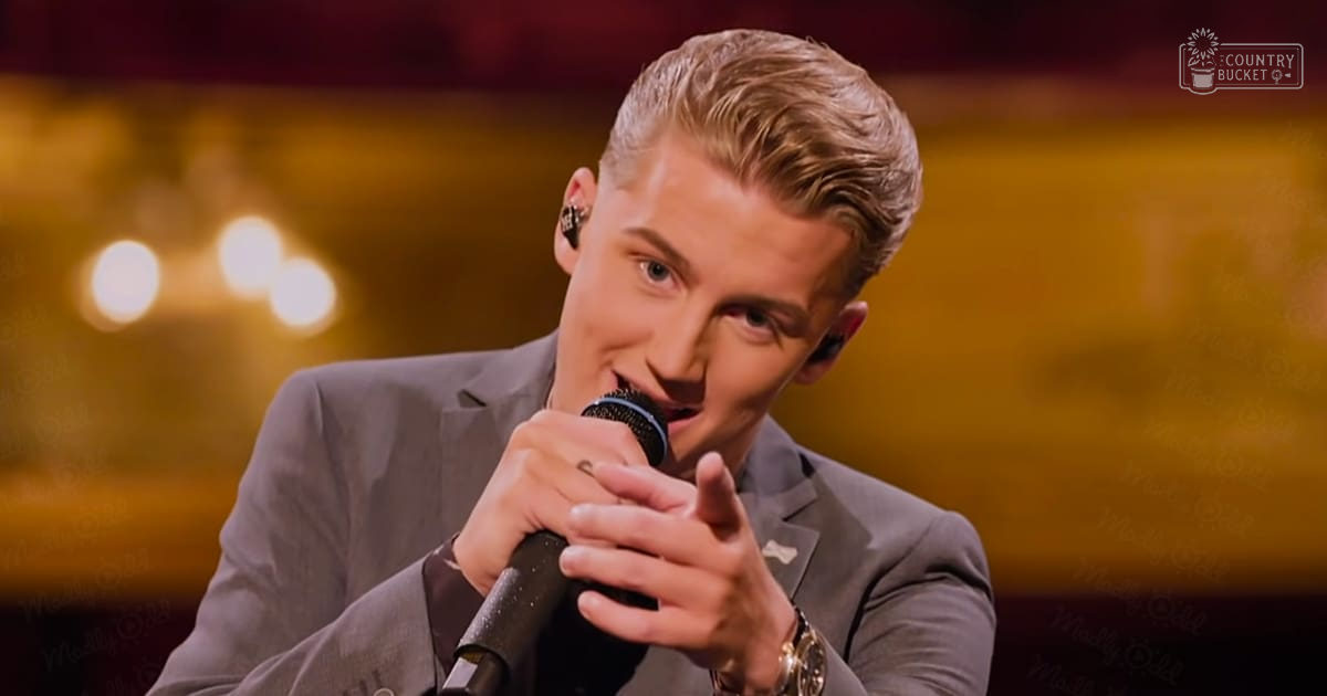 A young adult male singer wearing a gray suit jacket with subtle texture, performing on an indoor stage with blurred lights suggesting evening hours.