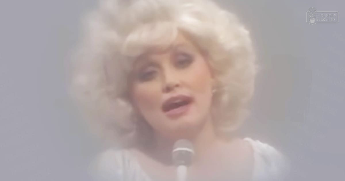 An adult female performer in a 1970s-style white ruffled sleeve top, holding a microphone on stage with soft diffused lighting and light fog effects, suggesting an indoor performance space.