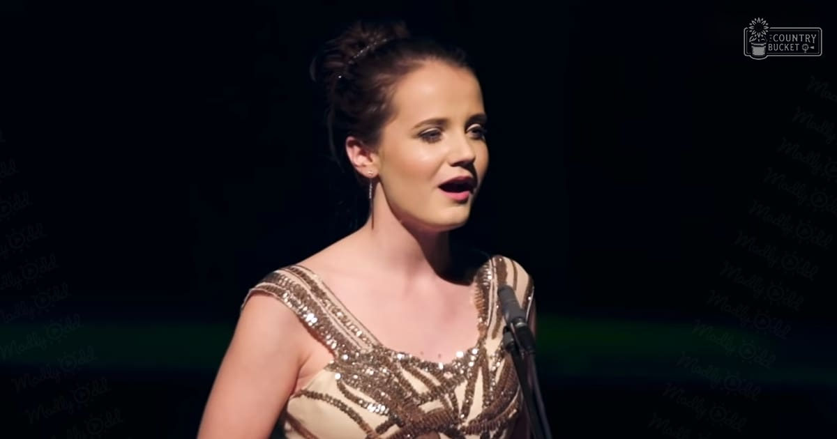 A young adult female in an off-the-shoulder dress with intricate beadwork and sequins performs on stage under dim lighting.