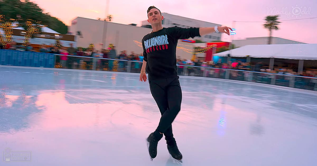 Young adult male in black 'Billionaire Boys Club' sweatshirt skating at an outdoor festive ice rink with pinkish dusk sky, social event setting.