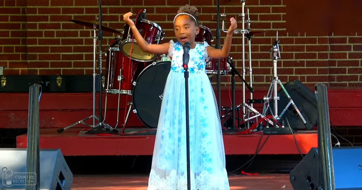 A young girl with dark skin tone, wearing a white dress adorned with blue floral patterns, standing on stage in front of a microphone stand during an outdoor event.