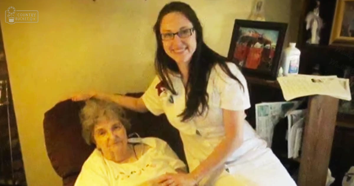 A young female caregiver in a white uniform with glasses assists an older adult woman seated in a dark wooden chair. The setting is indoors, likely a living room or bedroom, with warm lighting and framed pictures on the wall.