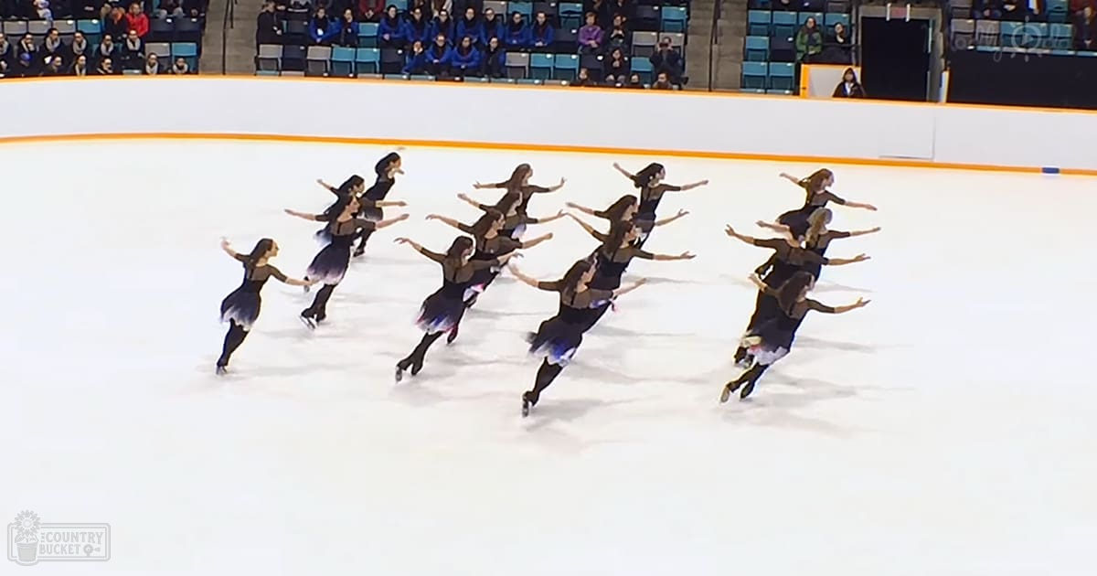 A synchronized skating team performs at an indoor ice rink with young female skaters wearing dark leotards and light blue skirts, surrounded by spectators seated on bleachers.