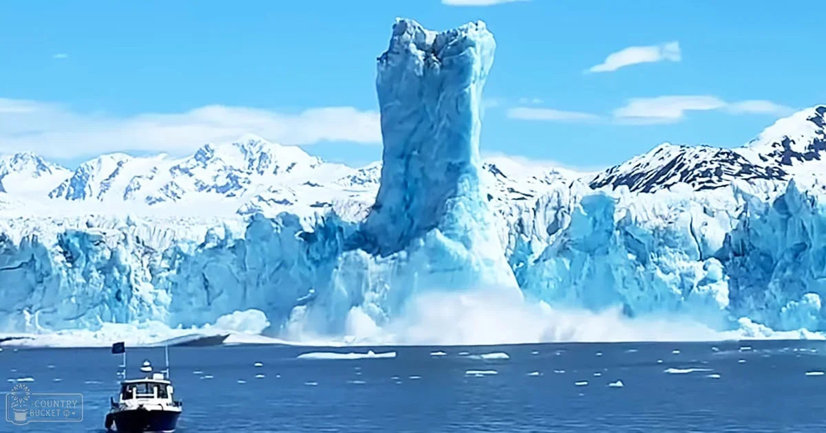 A large iceberg amidst snow-covered mountains, a small boat with a flag in the foreground on a clear day.