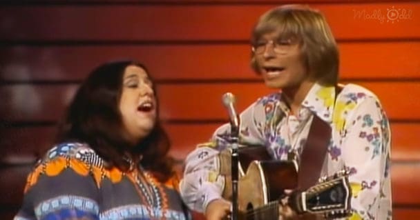 A musical performance scene with two adults, one female in a patterned top and male middle-aged wearing glasses. The setting appears to be an indoor performance space with wooden paneling and bright lighting.