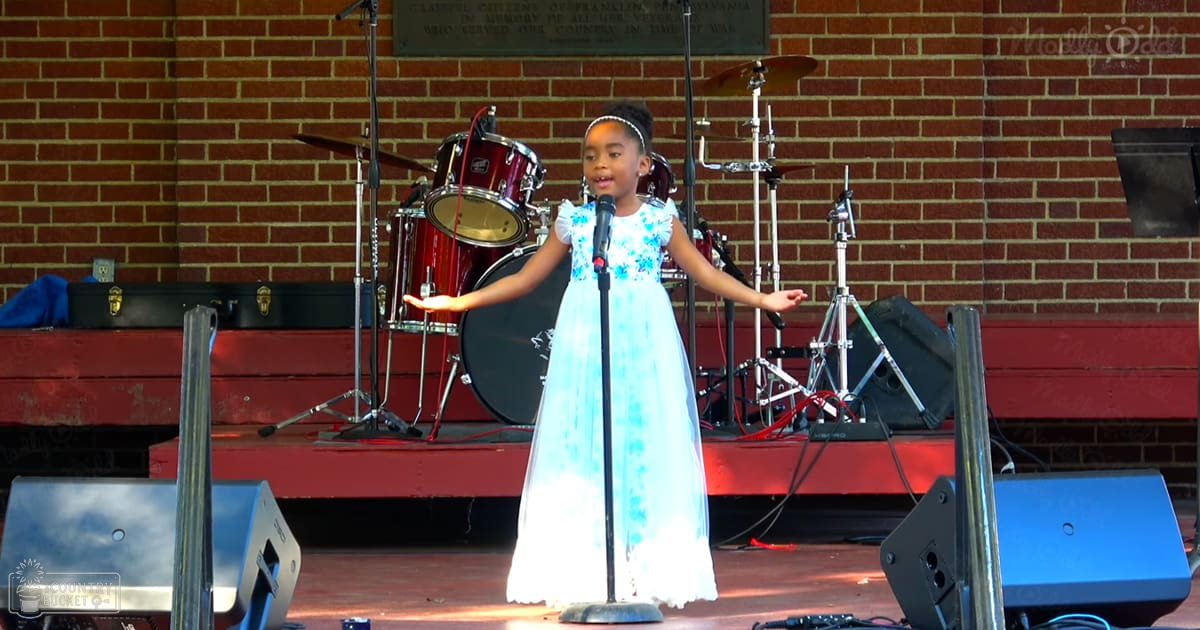 A child singing on a stage at an outdoor event, wearing a white dress with blue floral patterns.