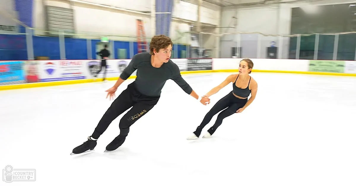 A young adult couple skating together in an indoor ice rink with advertisements visible in the background.