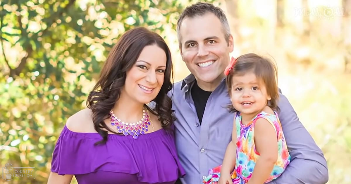 A family portrait taken outdoors with greenery and trees, with a young adult female in a purple top, an adult male in his 30s or 40s wearing a light gray shirt, and a toddler girl adorned with a red bow. The warm lighting suggests late afternoon.