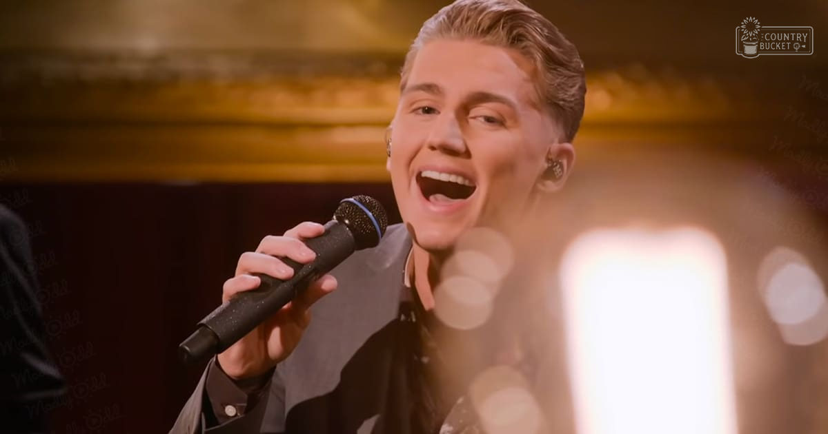 A young adult male singer in a black suit jacket with white lapels, performing into a microphone on stage under warm lighting.