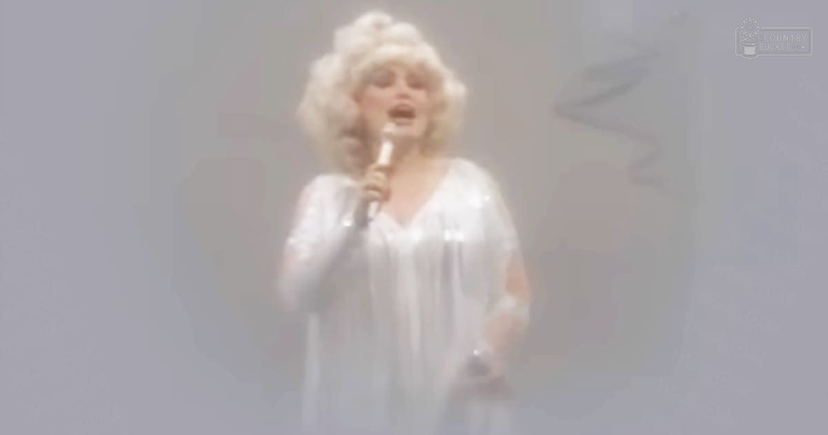An older adult female performer wearing a white, slightly sheer garment with ruffled details around the collar area at what appears to be a concert or performance setting.
