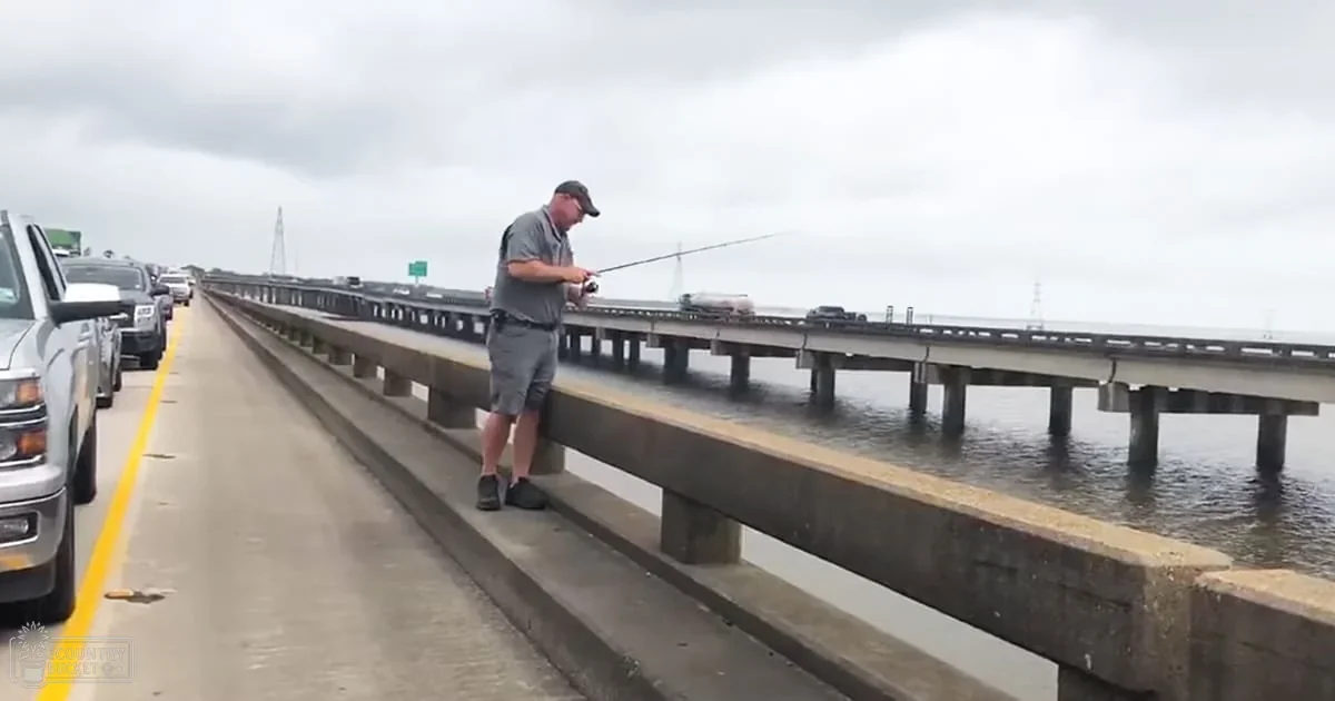A middle-aged man fishing from a concrete bridge over water near an elevated highway under cloudy skies.