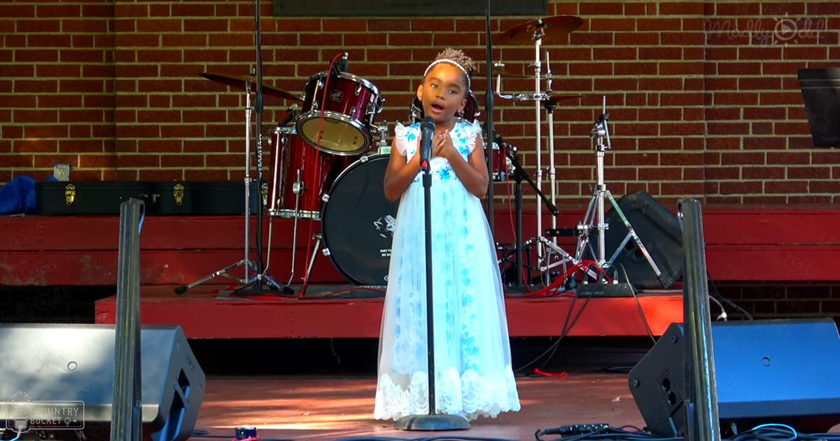 A young girl in a white tulle dress with blue floral patterns sings on stage at an outdoor event under daytime lighting.