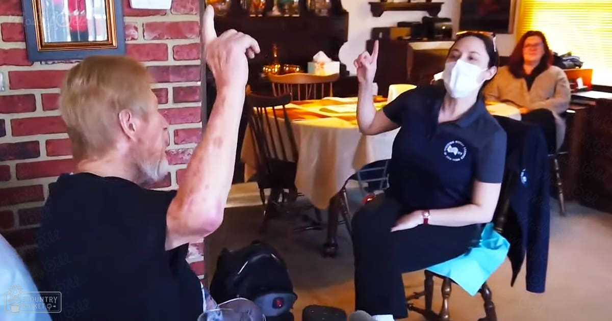 Middle-aged man and woman interacting in a home-like setting with dining furniture, brick wall art, and natural daylight.