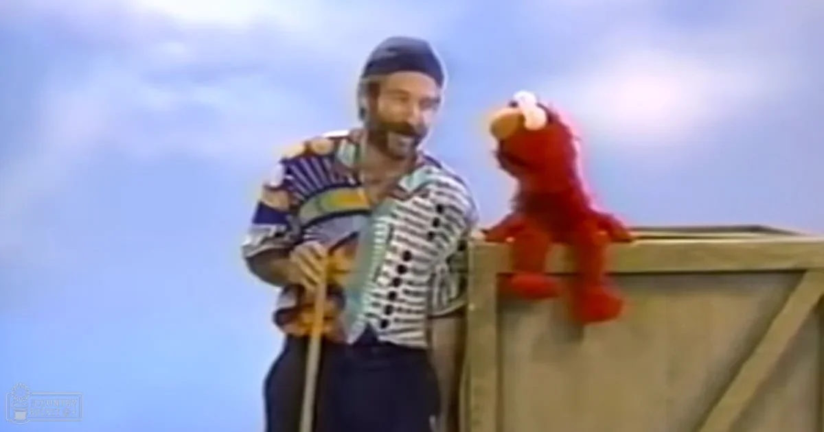 A Sesame Street Muppet named Elmo being held by an adult male puppeteer in a simple outdoor setting with blue skies and bright daylight.
