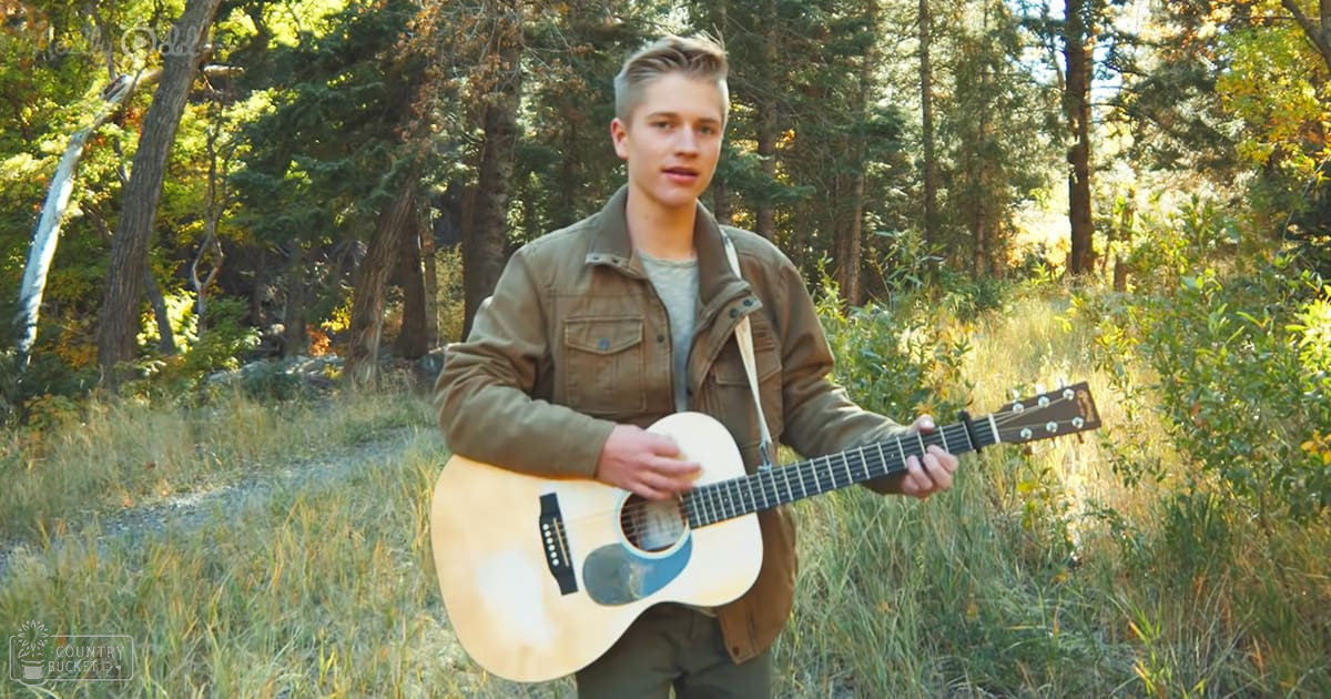 A teenager in a forested area, wearing a khaki jacket with two chest pockets, holding an acoustic guitar under warm afternoon light.
