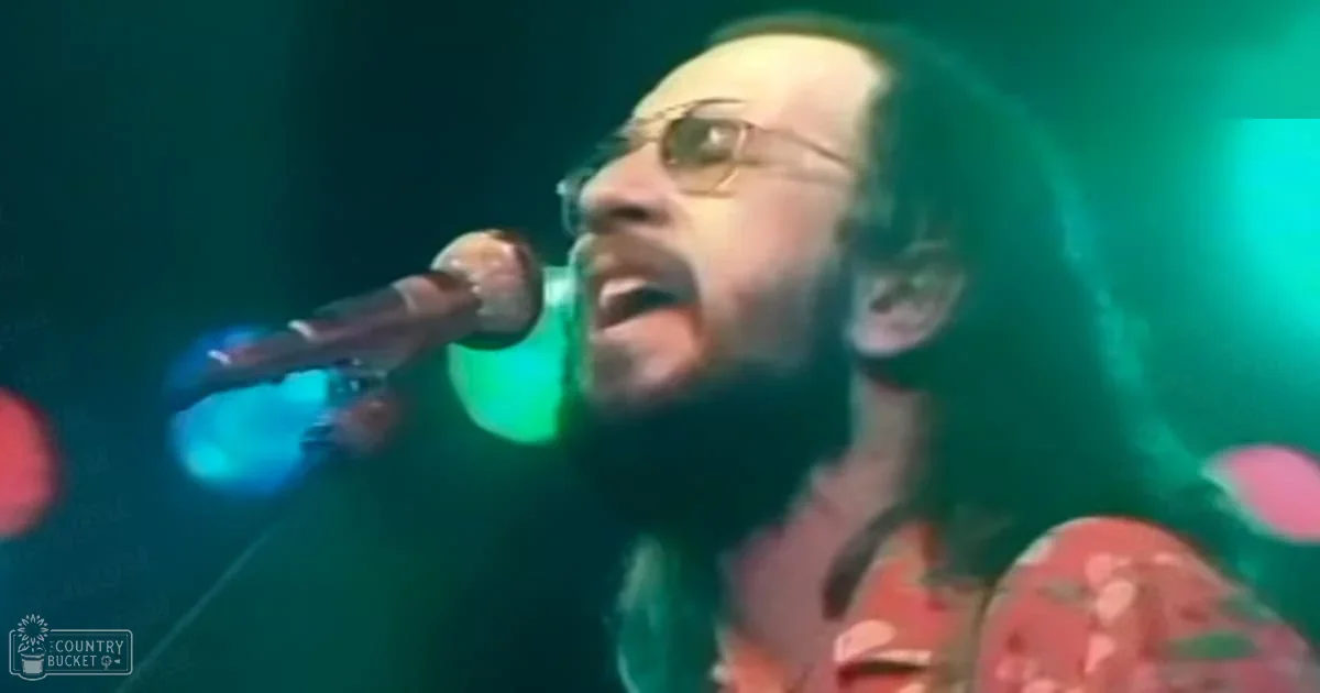 A middle-aged male singer wearing a floral patterned red and green 1970s or 80s era shirt with glasses, singing into a microphone at an indoor concert venue under stage lighting.