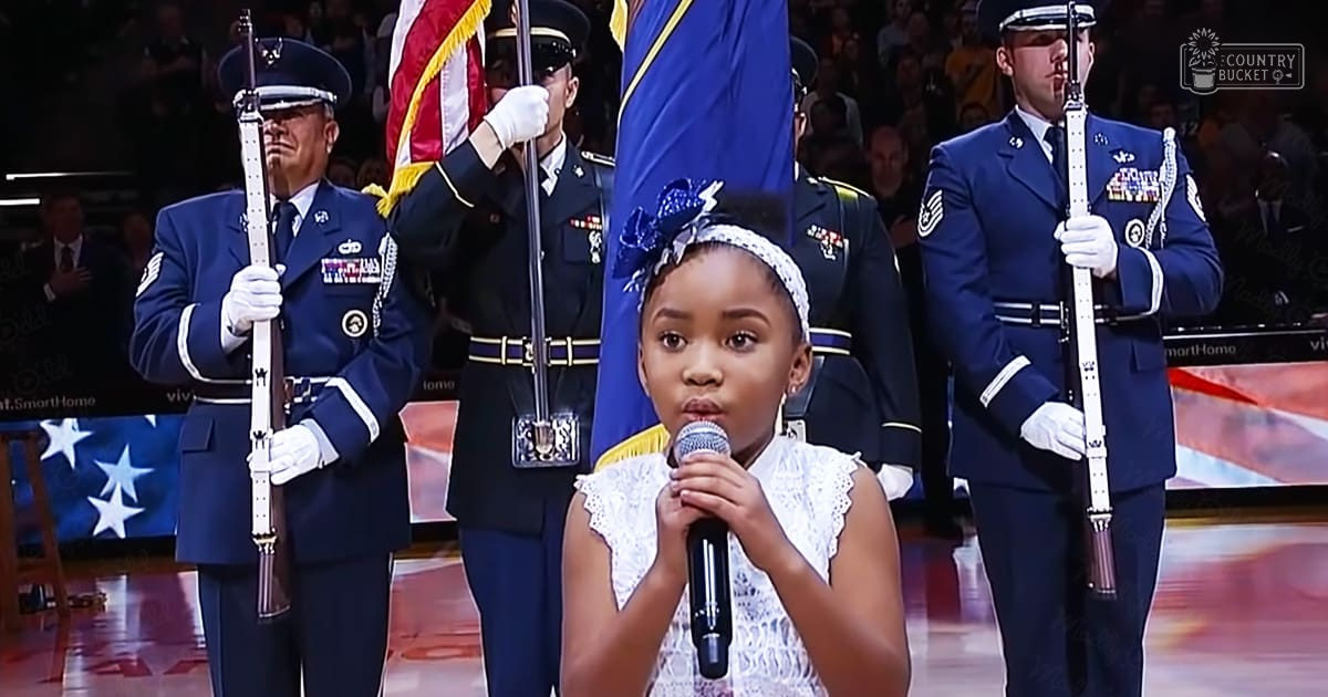 A young girl sings into a microphone at an indoor event with military personnel in uniform as part of the background.