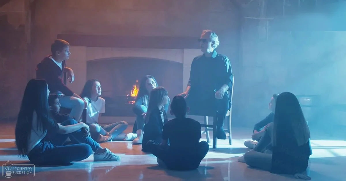A group of young adults in an indoor setting, possibly a classroom or meeting room, includes a child/teen wearing a white shirt and jeans, older adult male with glasses and dark blue shirt, and others in casual attire. The scene is dimly lit, suggesting evening or night, with stone walls and a polished wooden floor visible.