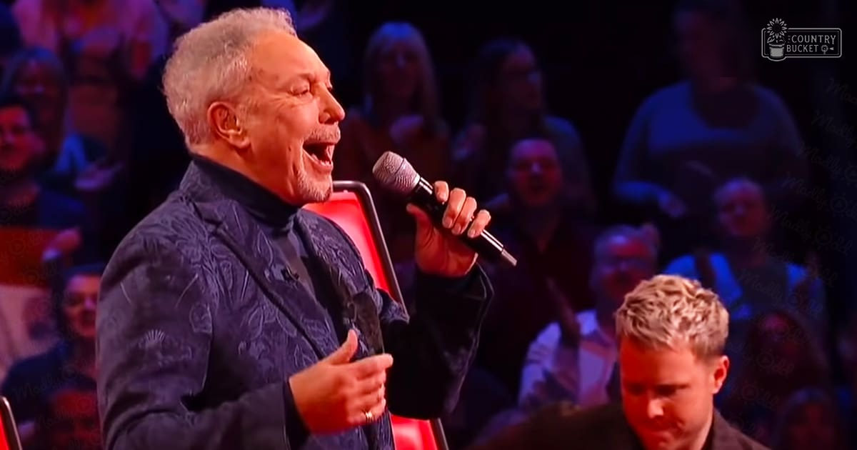 An older adult male singer in a television studio setting, wearing a patterned dark blue jacket with red chair in foreground.