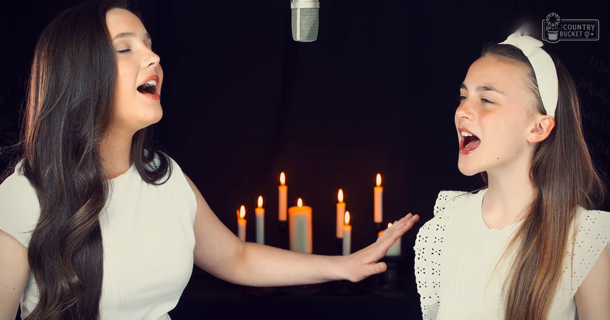 Two young adult females singing in a studio with dark walls and lit candles, wearing white tops; one with short sleeves and lace trim.