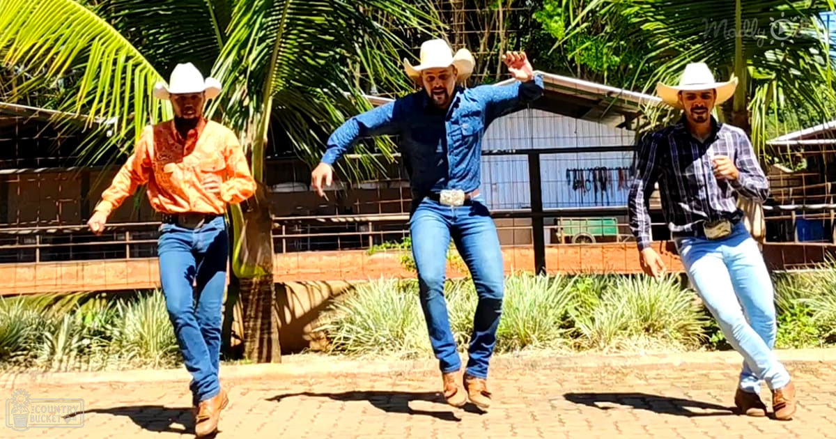 Three adult males dancing outdoors in a rural setting with palm trees and greenery under clear daytime skies.