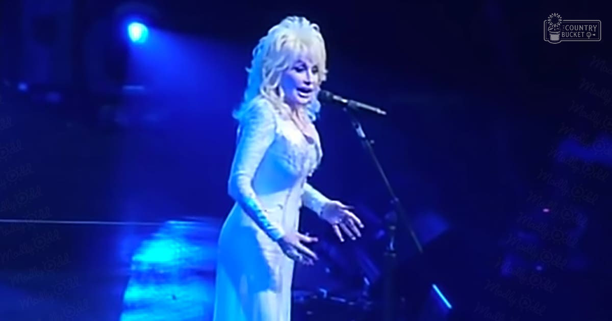 A female adult singer in an indoor concert venue, wearing a white long-sleeved vintage-style dress with a fitted bodice and flared skirt under dim blue stage lighting.