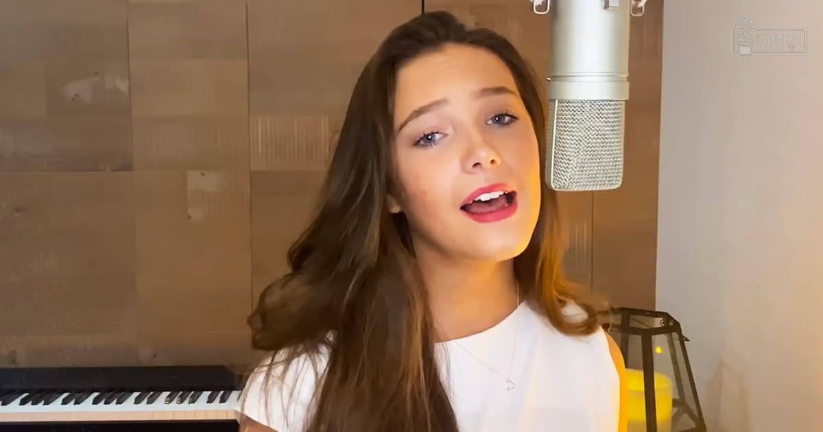 A young adult female in a relaxed white t-shirt, delicate necklace, recording studio with piano keyboard, microphone stand, and decorative lantern.