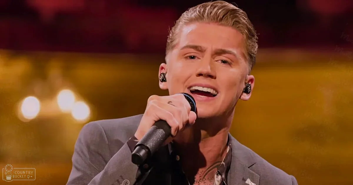 An adult male singer in a gray suit jacket with black shirt, performing on an indoor stage illuminated by warm yellow lights.