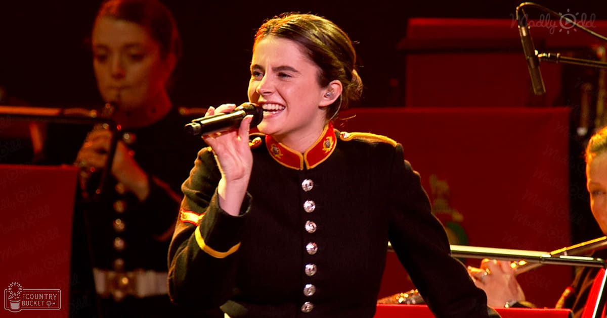 A young adult female sings into a microphone, wearing a formal military-style uniform with red collar and gold buttons, set against an indoor stage backdrop under red lighting.