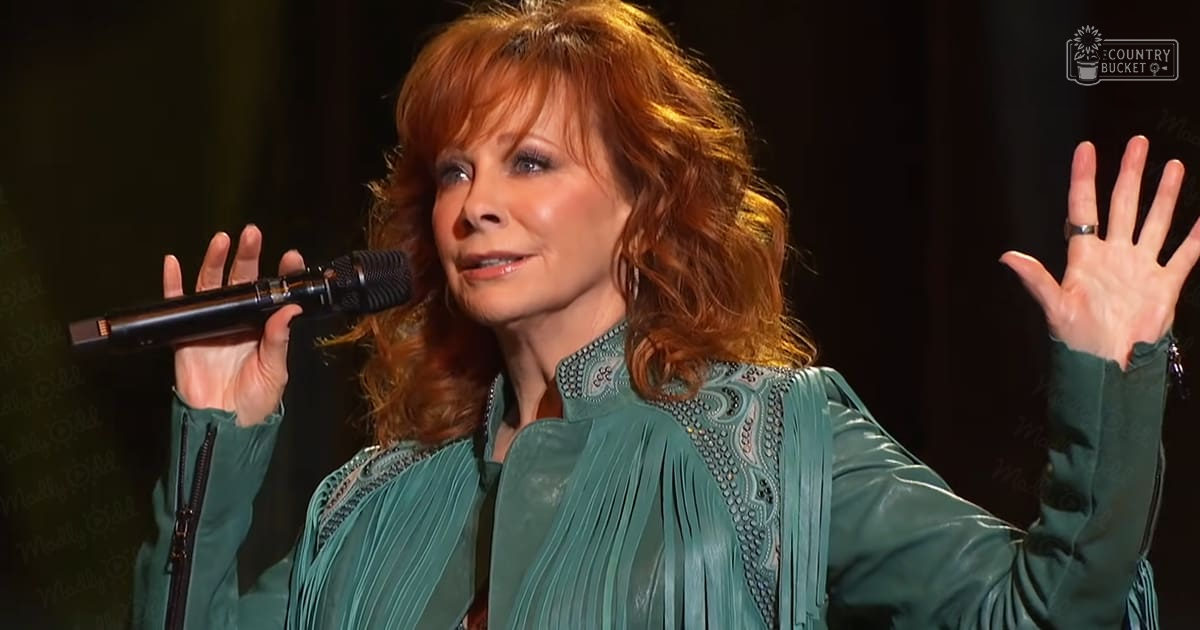 A middle-aged female performer wearing a teal, intricately embroidered and pleated top on stage during what appears to be a concert.