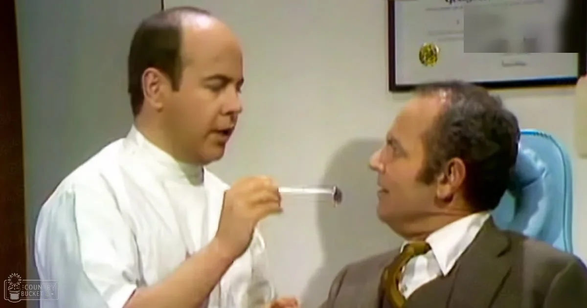 A middle-aged male medical professional examines a middle-aged to older adult male patient wearing a dark suit and patterned tie, seated on a blue chair in an indoor office setting with neutral-colored walls and a framed certificate.