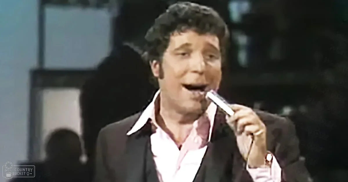 Adult male singer wearing dark suit over pink shirt, holding cigarette, stage setting with blurred figures in background.