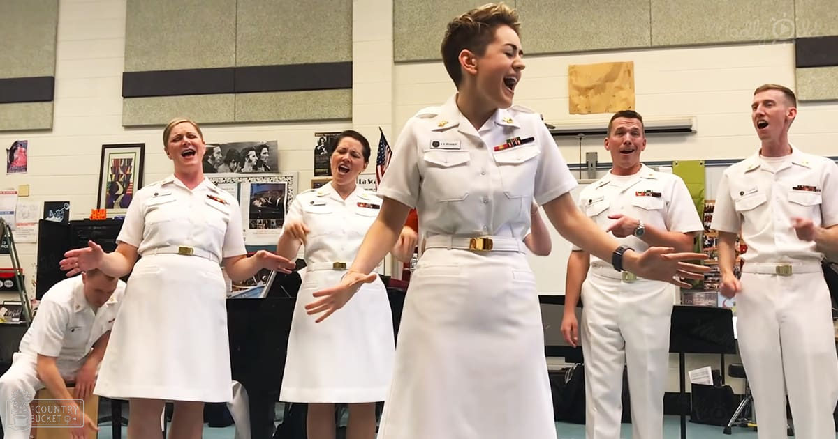 A group of young adult females and males in white naval uniforms singing indoors at a military base or similar institution. The scene includes acoustic panels, music stands, and posters on the wall.