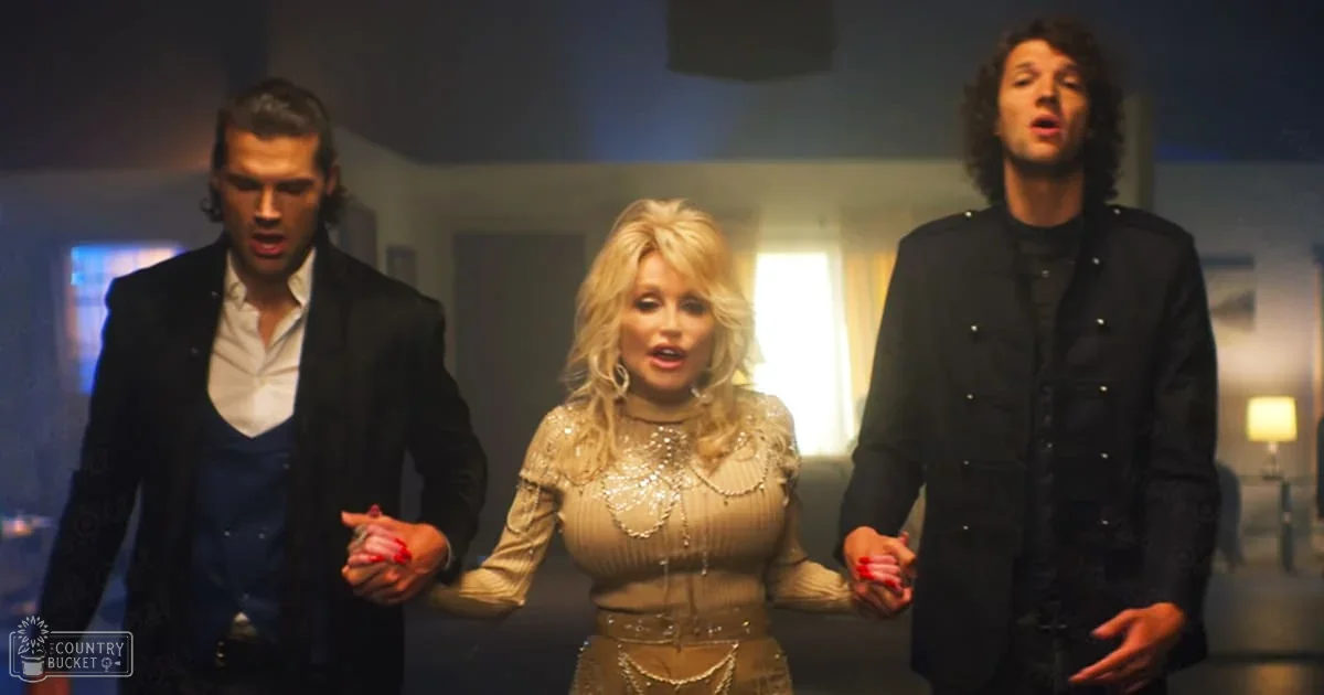 A middle-aged woman in an elegant beige top with intricate embellishments stands between two young adult men dressed formally in blazers and double-breasted jackets. The setting appears to be a modern indoor interior, possibly at an event or gathering.