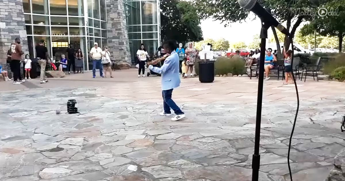 An outdoor performance with performers in casual summer attire near modern buildings with glass windows and green trees.