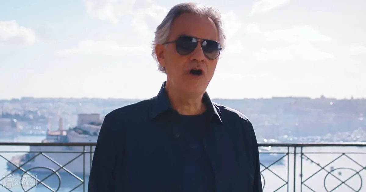 An older adult male stands on a balcony or terrace, overlooking a body of water with a ship docked nearby; partly cloudy sky suggests midday in spring or summer.