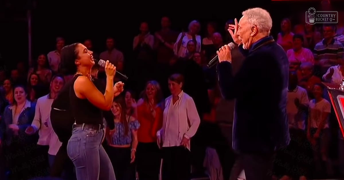 A musical performance scene in an indoor venue, with two performers singing into microphones on stage while an audience watches under artificial light.
