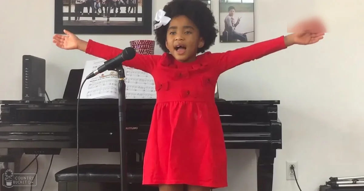 A young child wearing a red dress with a bow, singing into a microphone indoors among framed photos and artwork.