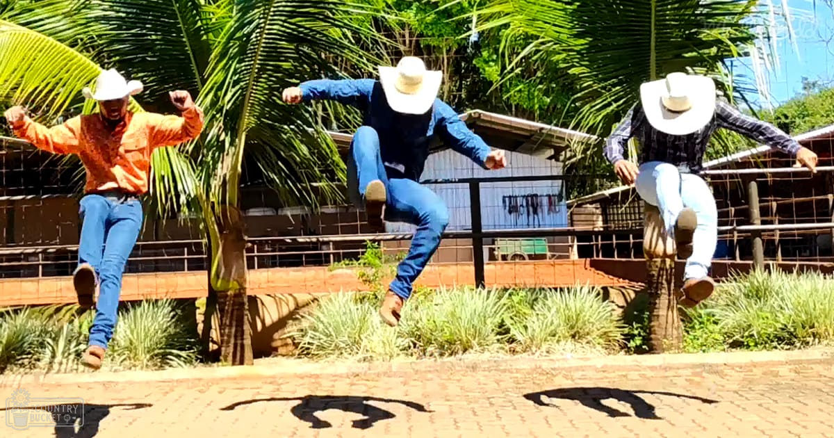Three adult males in cowboy attire performing a synchronized jump on an outdoor farm or ranch with greenery and a clear sky.