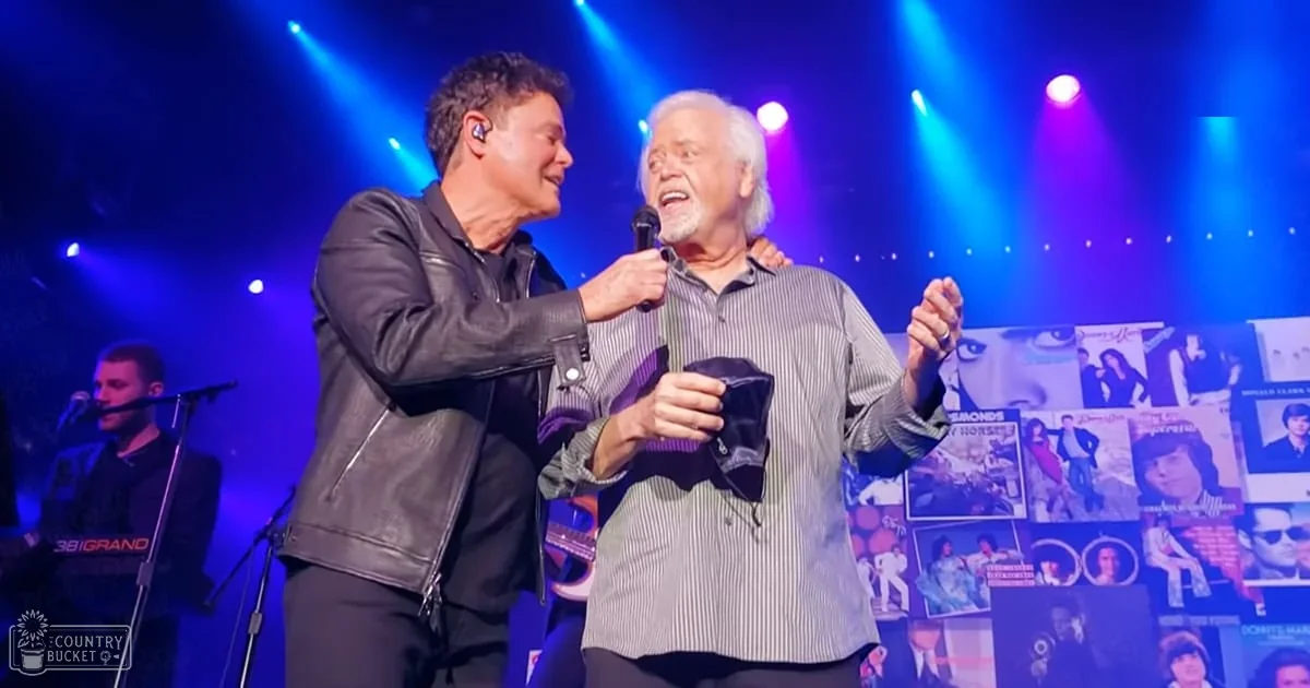 Two adult males performing on stage, one wearing a black leather jacket and the other a striped button-up shirt with a microphone stand visible behind them. The backdrop includes images of people and text.