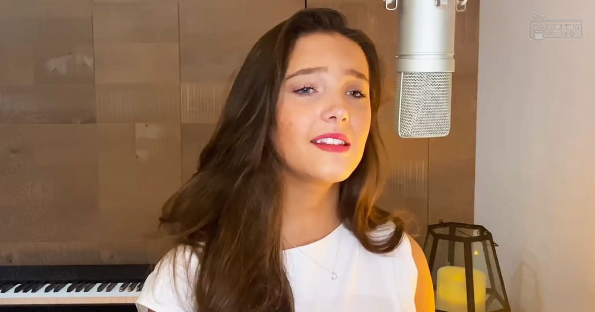 A young adult female wearing a white t-shirt in an indoor recording studio setting with warm lighting and soundproofing panels.