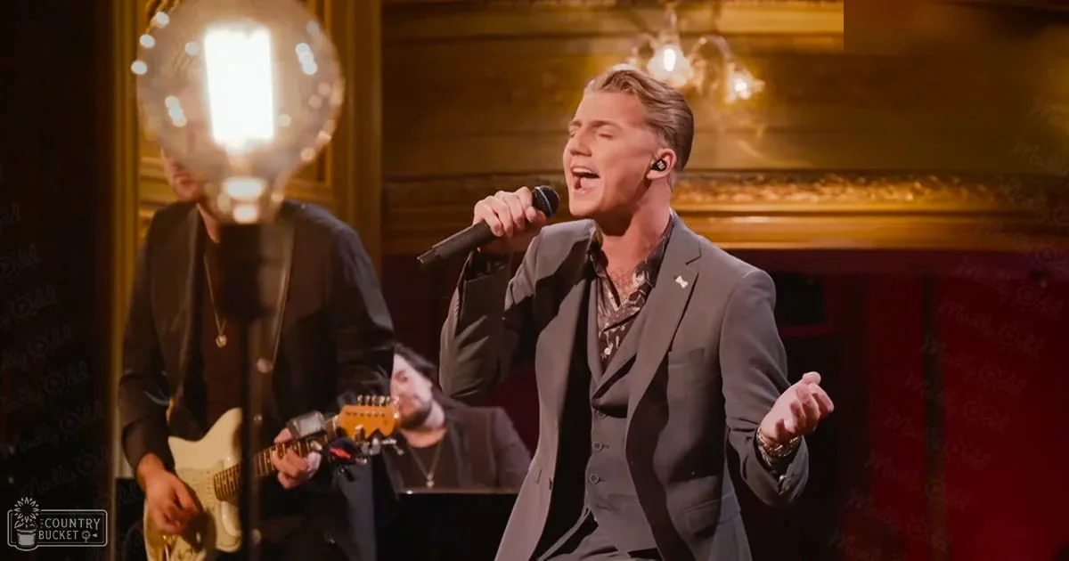 A young adult male singer performing in an indoor venue wearing a dark gray suit with a classic collar and patterned tie.