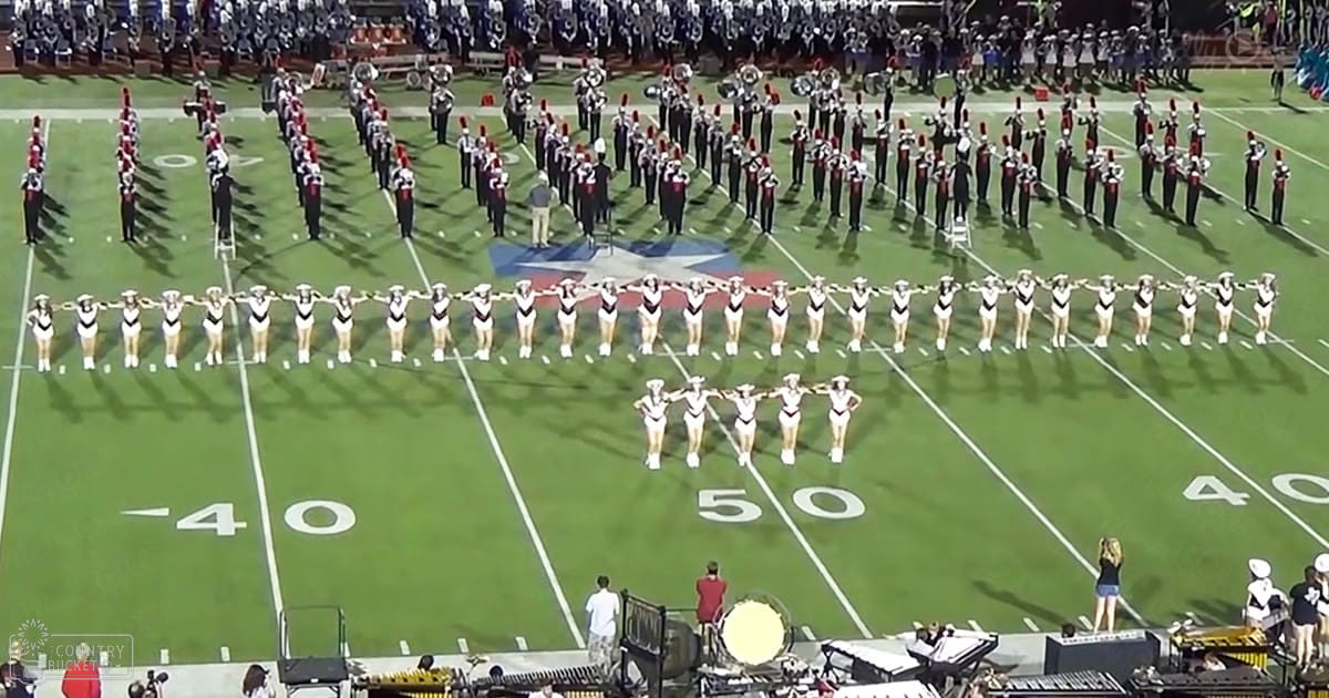 A marching band performance at a football stadium during halftime, with cheerleaders in white and black uniforms with gold accents and young adult to middle-aged male marching band members in red and white uniforms with black pants and red hats.