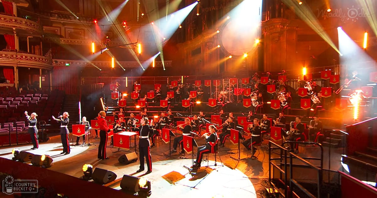 Military band performing at an ornate concert hall with bright stage lighting and rows of empty seats, red uniforms with gold emblems hanging behind musicians.