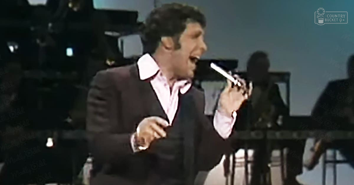 A male adult singer in a dark suit jacket over a white shirt with red stripes and a black tie performs into a microphone on stage, likely in a mid-20th century studio setting surrounded by musicians seated at a piano.