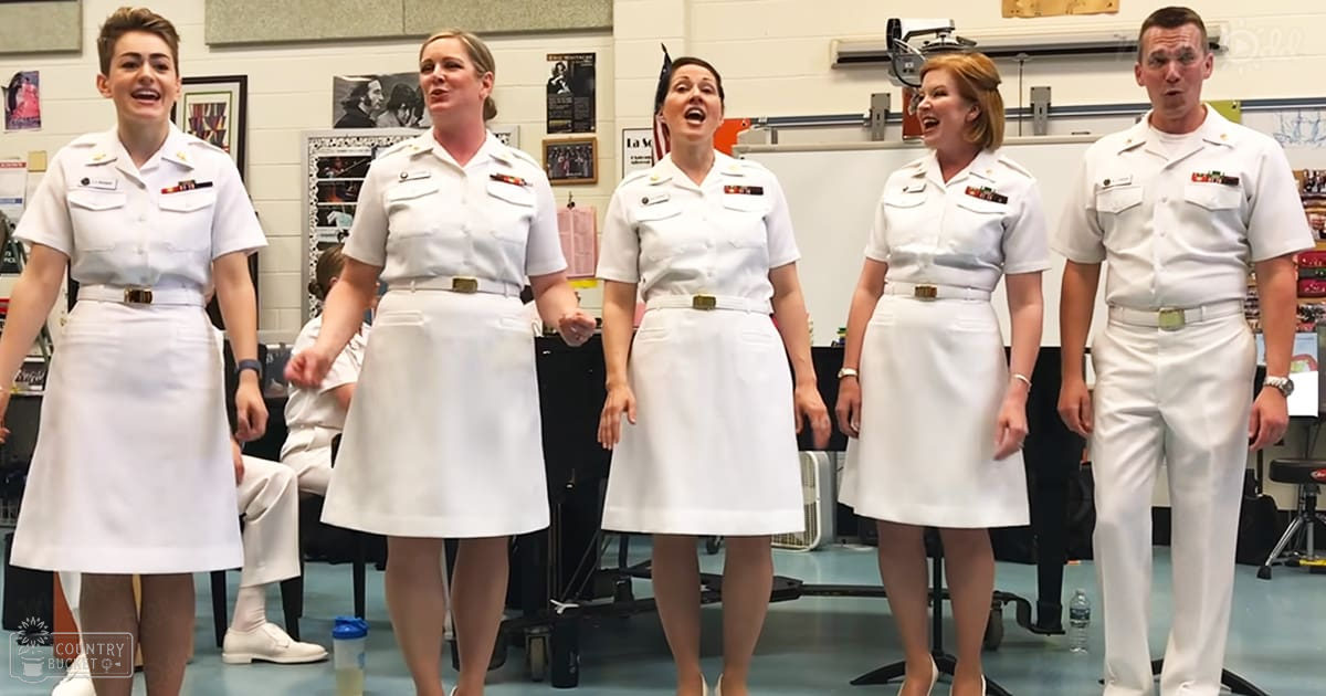 A group of young adult females and a middle-aged female in military-style white shirts, possibly at an educational facility. The setting is indoors with walls adorned with educational posters.