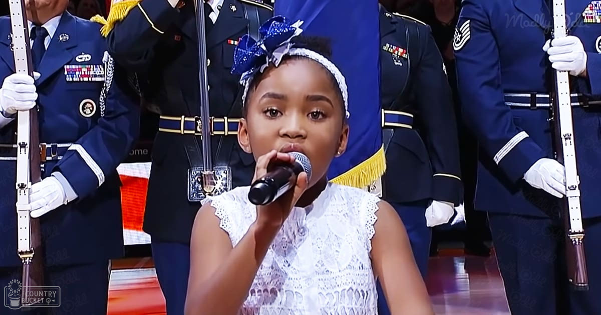 A young girl in a white lace dress sings into a microphone at an event with military personnel in uniform. The setting appears to be an indoor military ceremony or formal occasion.