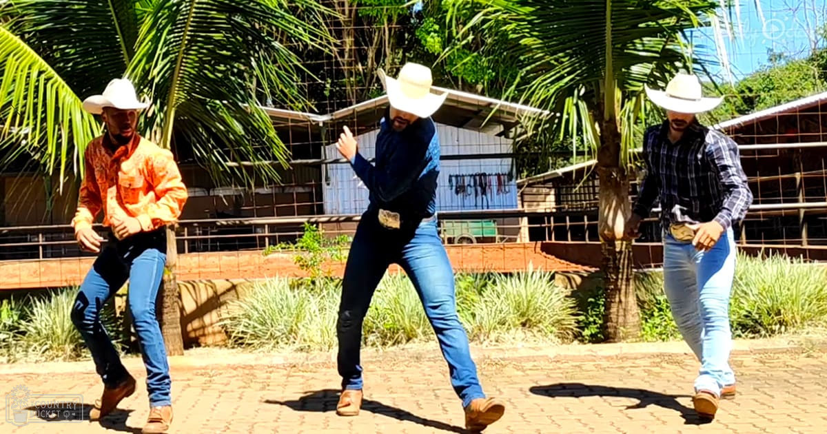 Three adults dancing outdoors in a rural setting with palm trees and farm structures, wearing cowboy attire.