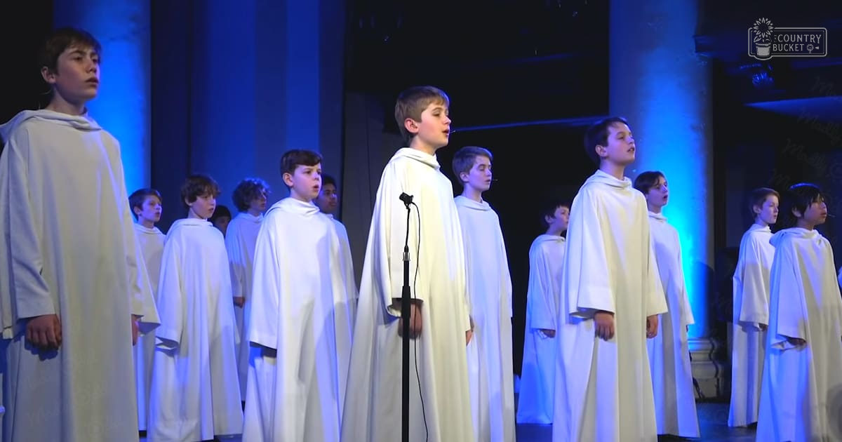 A children's choir performing indoors on a stage under blue lighting, with white robes and curtains in the background.