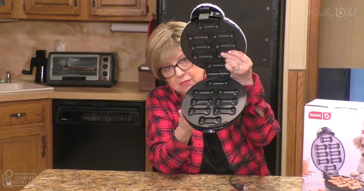 A middle-aged adult female in a kitchen setting, wearing a red plaid shirt with long sleeves and glasses, holding an open waffle maker.
